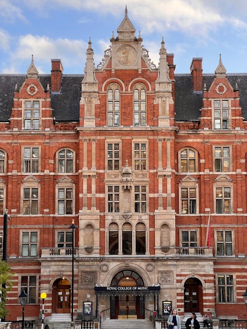 A detailed view of the exterior of a historic red brick building with ornate stone features, large arched windows, and decorative gables, located on Exhibition Road in South Kensington. The building h